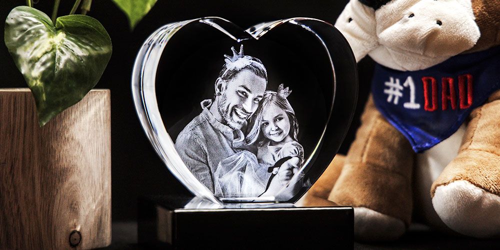 Heart-shaped crystal on a base, engraved with a father and daughter smiling together, with a plush toy and houseplant nearby.