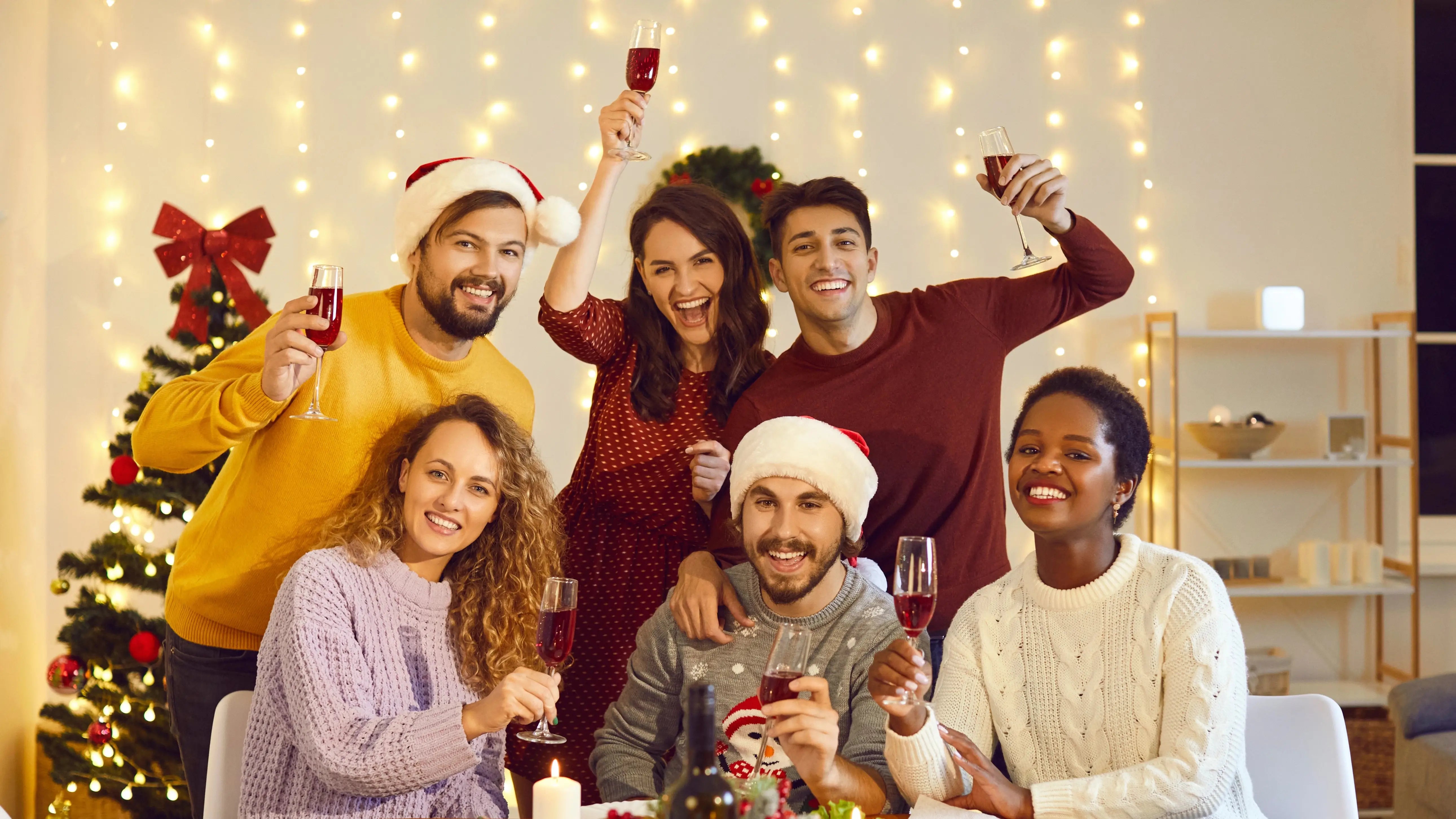 Friends happily toasting and celebrating christmas together in a dining room with christmas lights and a christmas tree behind
