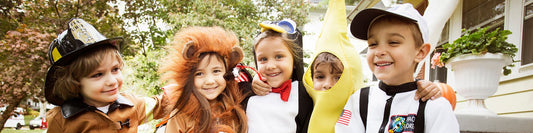 A group of five young children in fun Halloween costumes — a firefighter, lion, penguin, banana, and astronaut — smiling together outdoors during fall.