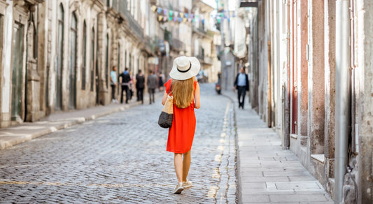 A woman in a red dress and white hat walks alone down a charming cobblestone street lined with old stone buildings and a few p