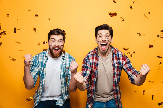 Two excited men celebrating with confetti falling against an orange background, showing their team spirit with face paint.