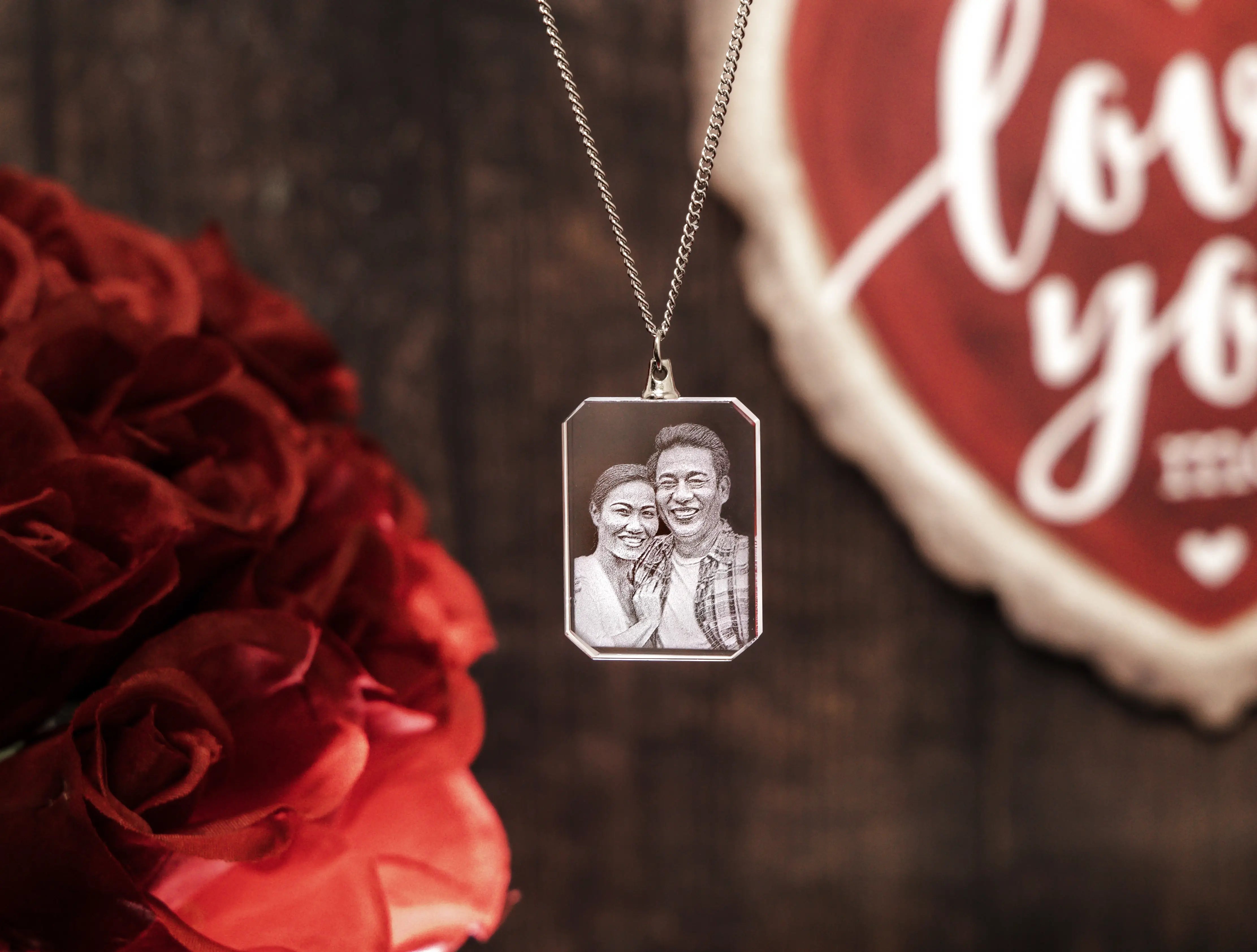 Close-up of a rectangular crystal necklace engraved with a joyful couple’s portrait, hanging in front of red roses and a rusti