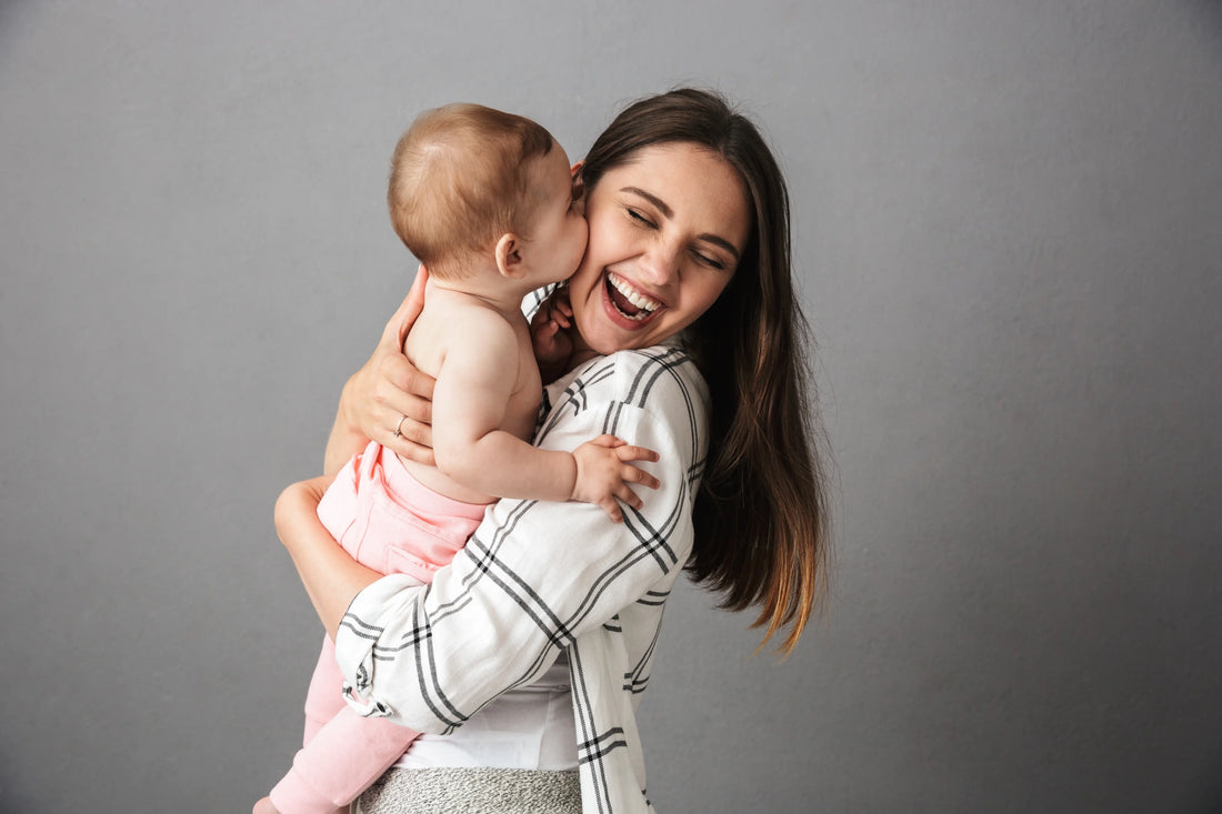 Smiling young mother holding a baby who is playfully kissing her cheek in front of a gray background.