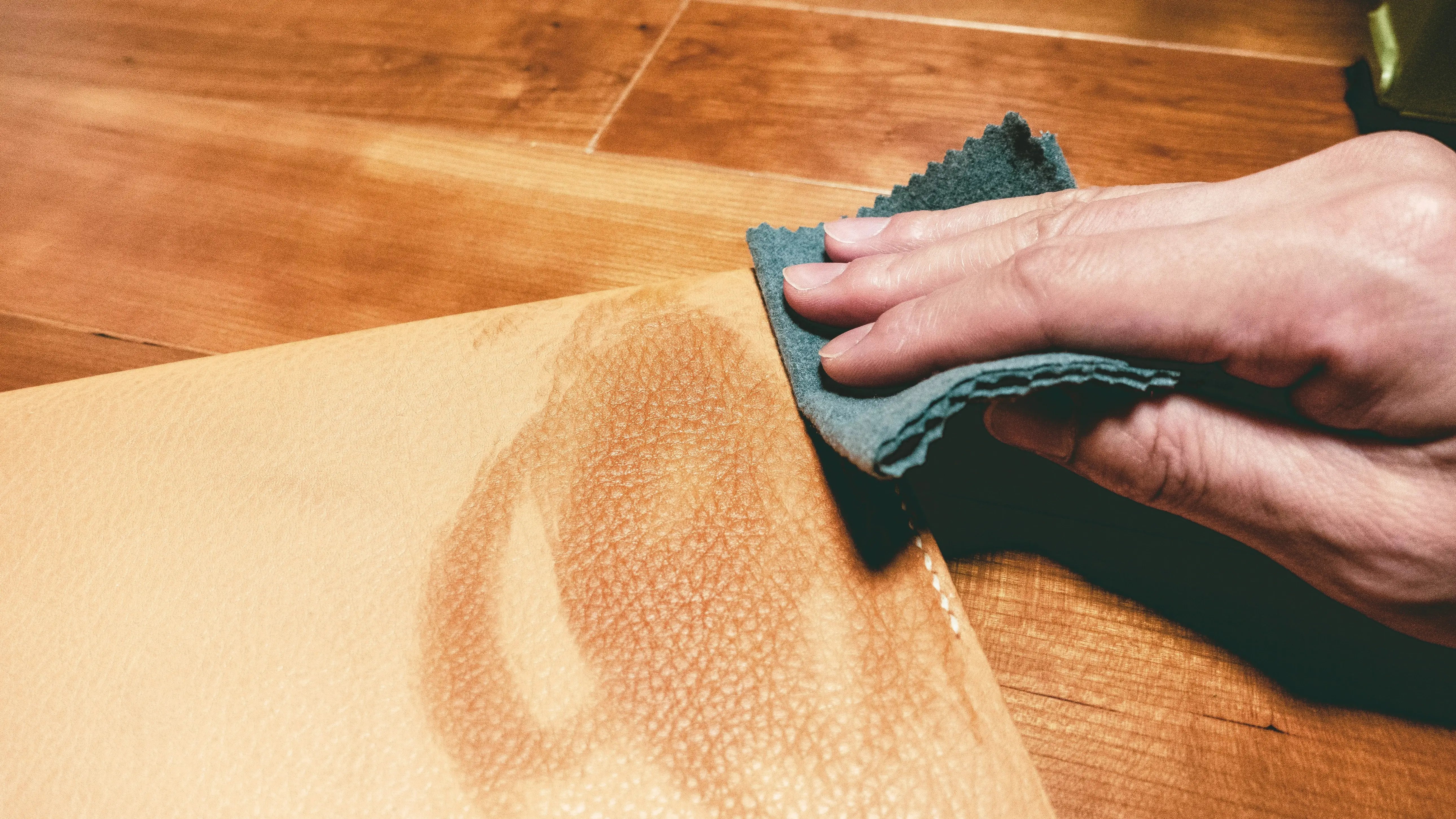 Hand using a cloth to clean a tan leather surface, demonstrating leather maintenance on a wooden table.