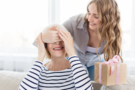 A cheerful daughter surprises her smiling mother by covering her eyes and holding a wrapped gift with a pink ribbon, celebrati