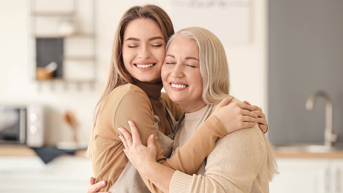A young woman and an older woman smiling with eyes closed while hugging warmly in a cozy, softly lit kitchen.