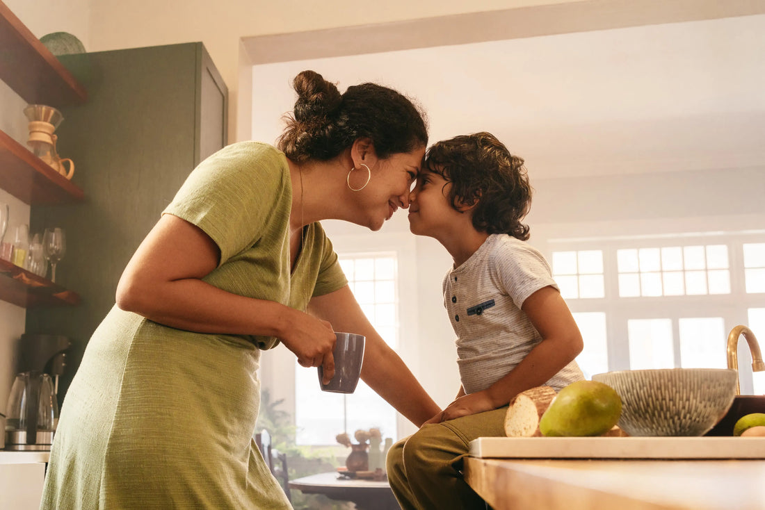 A sweet moment between a mother and her young son as they touch foreheads and smile at each other in a warmly lit kitchen.