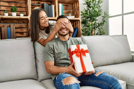 Smiling woman covering her partner’s eyes as he holds a white gift box with a red ribbon on a cozy couch.