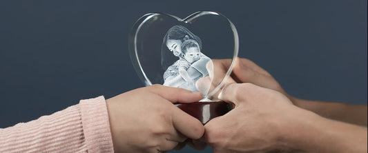 Close-up of two pairs of hands holding a heart-shaped crystal featuring an engraved image of a mother hugging her baby, symbol