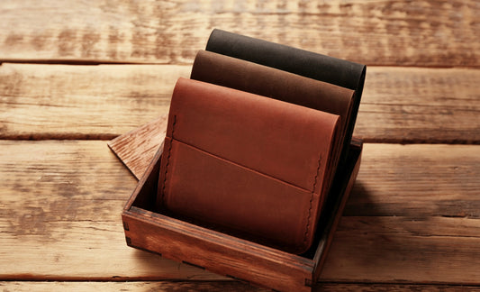 Three leather wallets in different shades—black, brown, and tan—neatly stacked inside a rustic wooden box on a wooden surface.