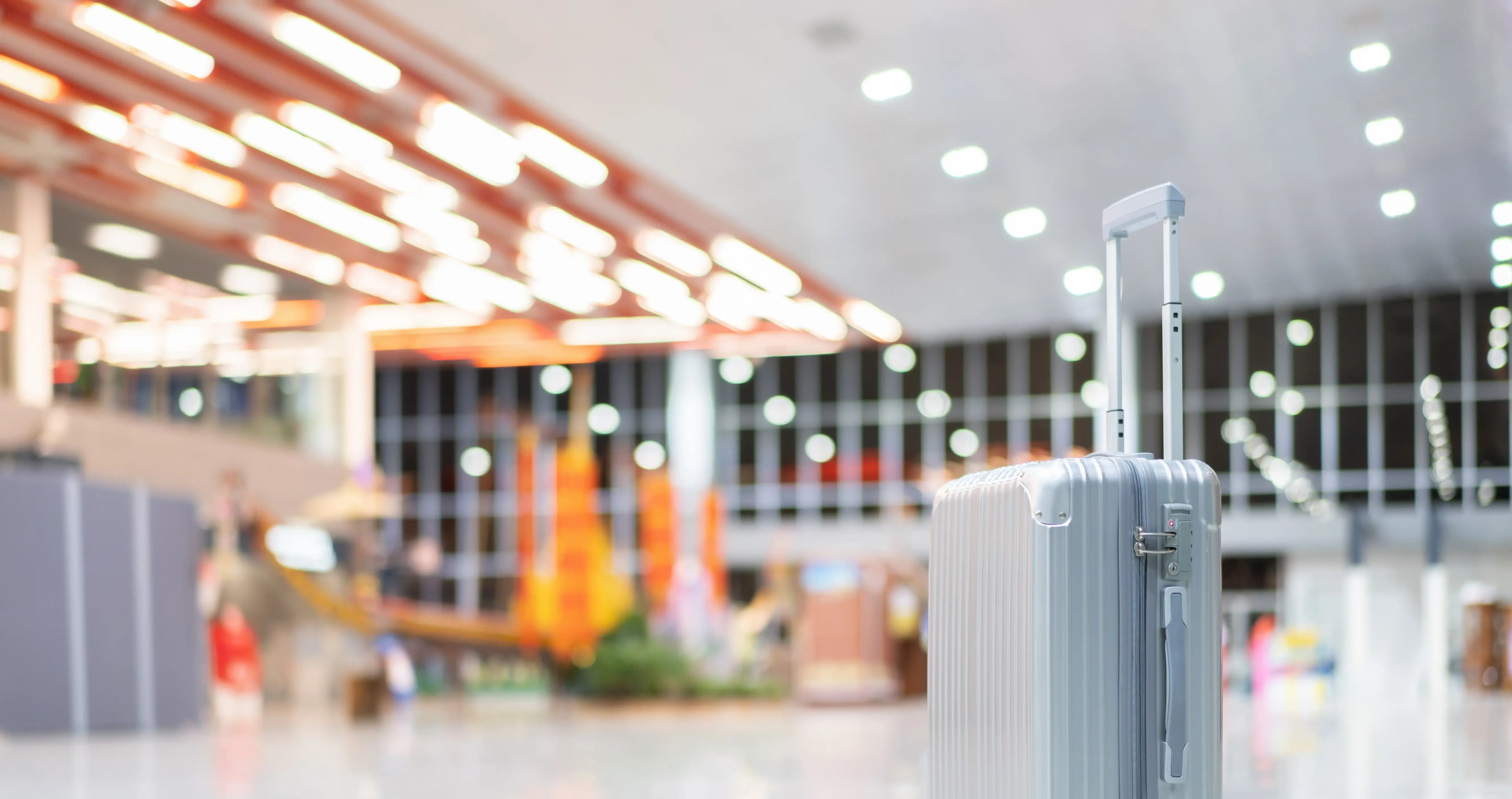 A silver hard-shell suitcase with extended handle standing alone in a brightly lit, modern airport terminal.