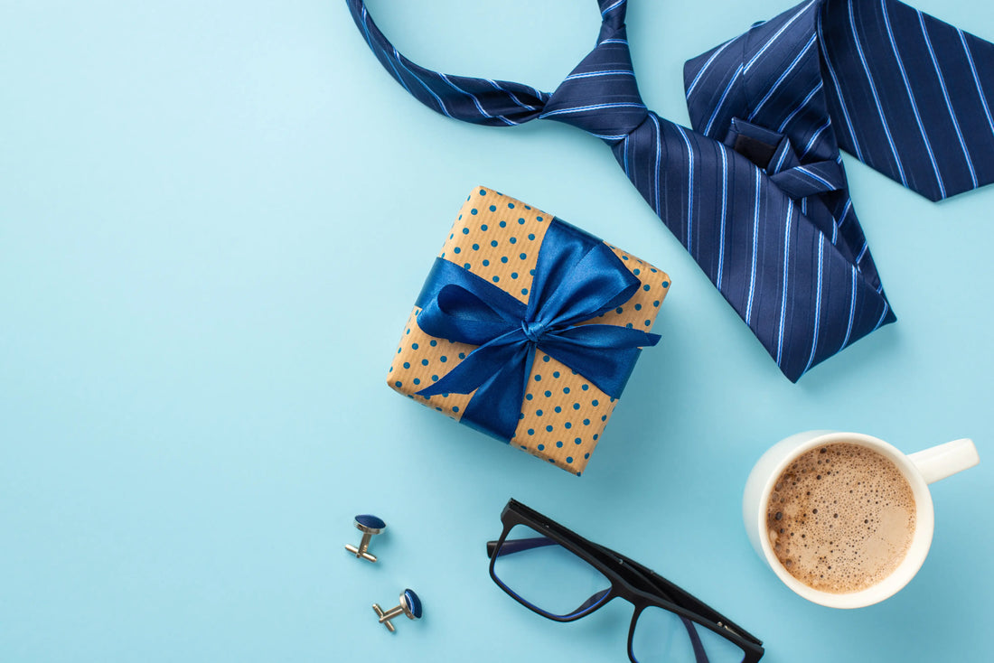 Flat lay of a wrapped gift box with blue ribbon, a striped necktie, a pair of cufflinks, eyeglasses, and a coffee mug on a lig