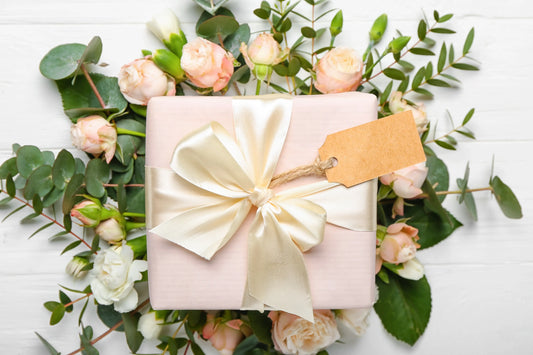 A pastel pink gift box with an elegant cream satin ribbon and a blank tag, surrounded by soft pink roses and green eucalyptus 