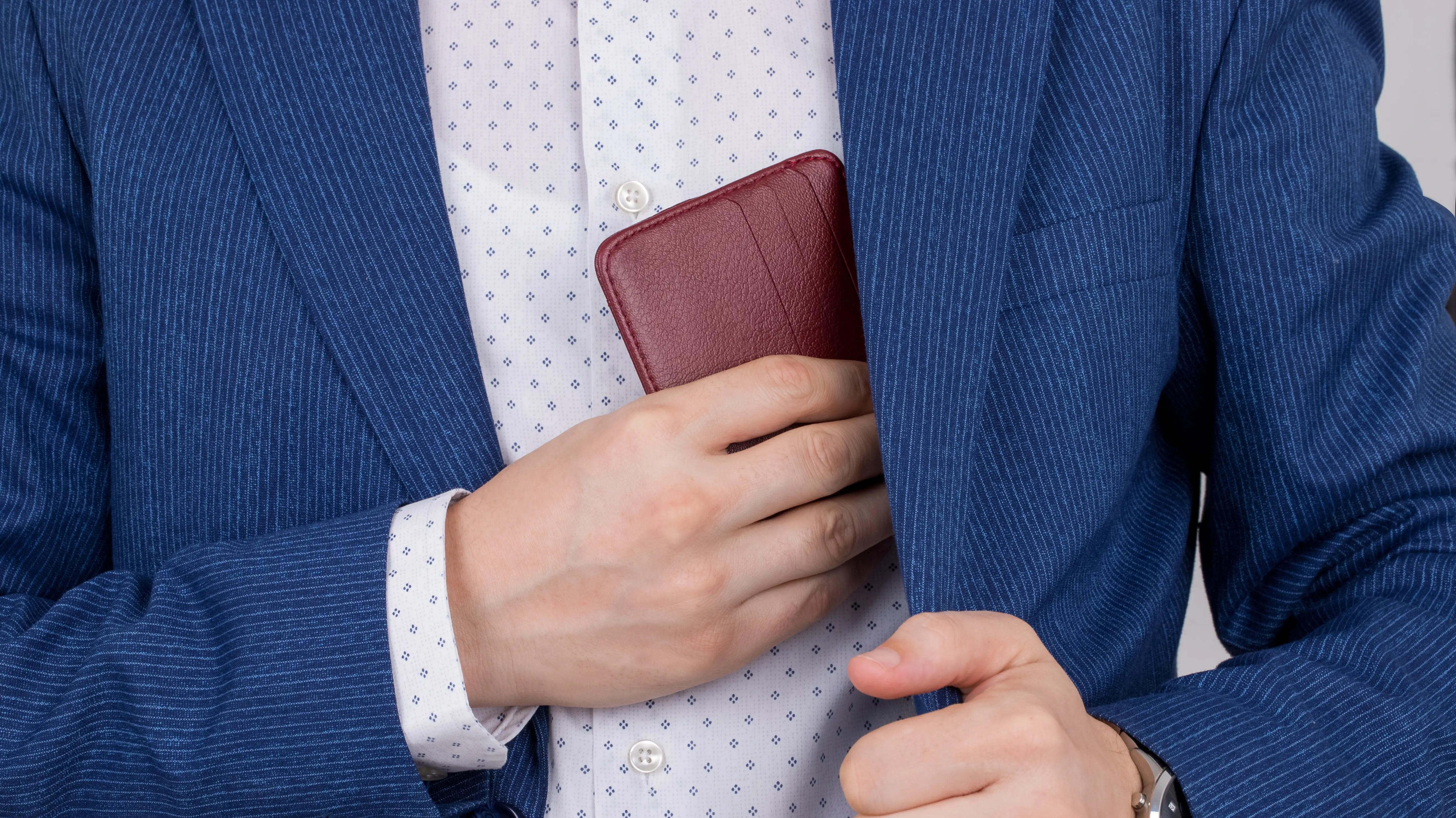 Man in a blue pinstripe suit placing a red leather wallet into the inner pocket of his blazer.
