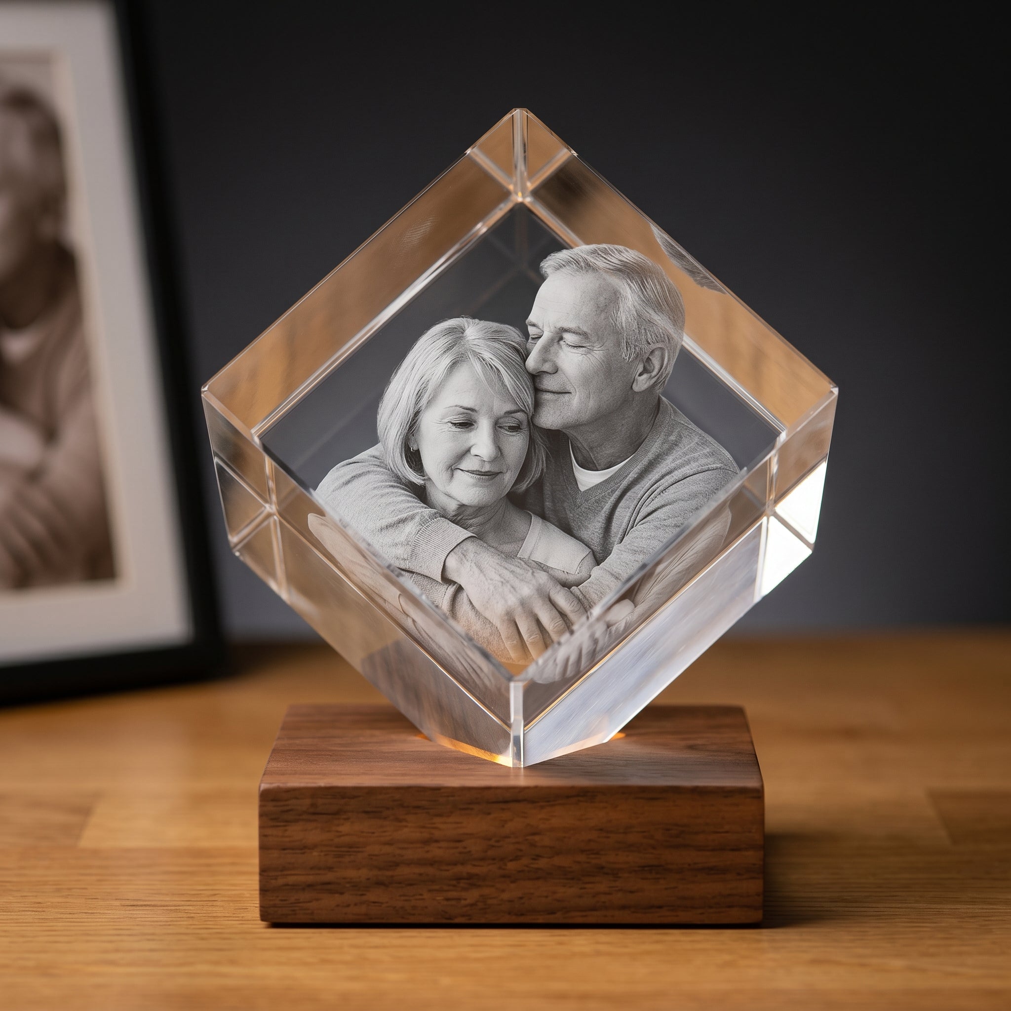 A diamond-oriented K9 crystal cube on a rosewood base shows an engraved older couple in a quiet embrace, with a bokeh framed print at the edge.