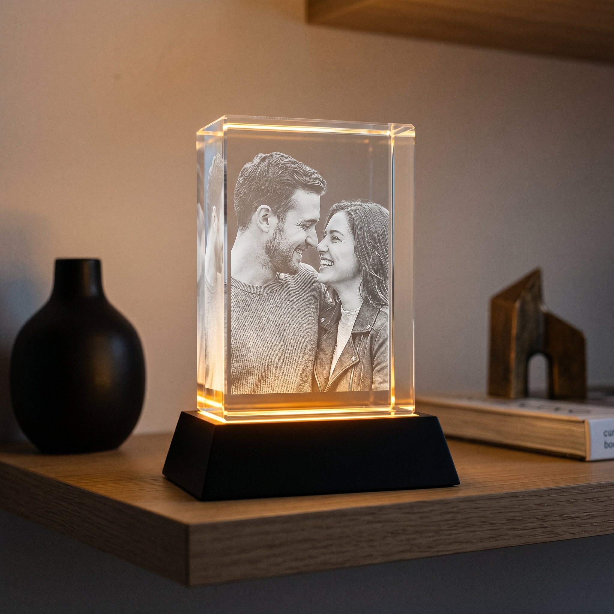 BM rectangle 3D crystal of couple looking at each other, black premium base showing prominent warm LED top-joint glow, contemporary floating wall shelf with matte vase and bronze sculpture